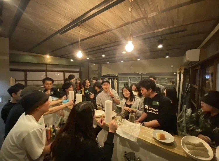 麦酒店のカウンターに、メンバーと友達がぎゅっと集まって乾杯している様子
