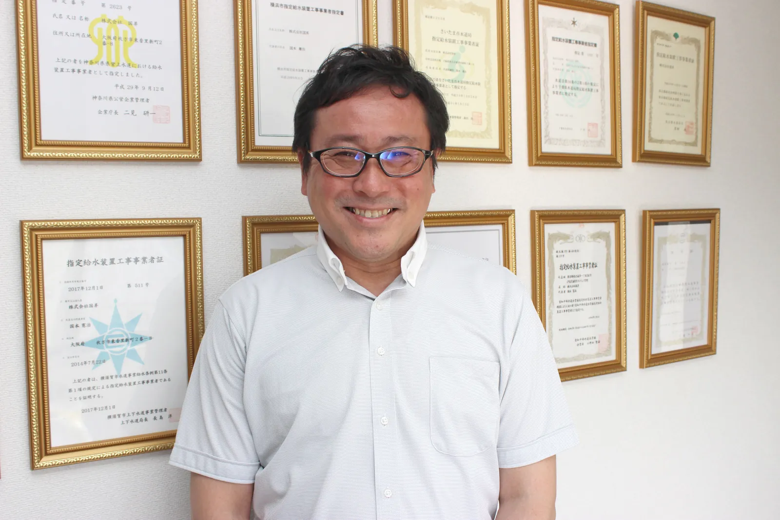 表彰状が並ぶ壁の前で笑う国本社長